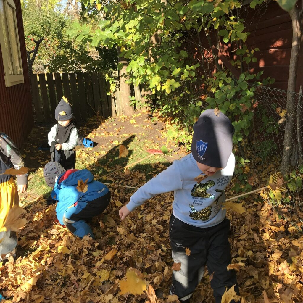 Barn leker bland höstlöv på Elefantens förskolegård, omgiven av träd och röda träbyggnader.