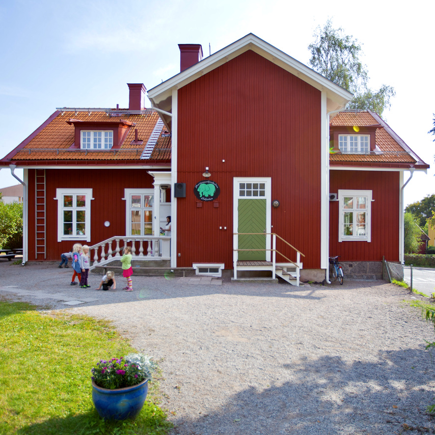Montessoriförskolan Elefantens röda trähus med grön entrédörr och barn som leker på gården.